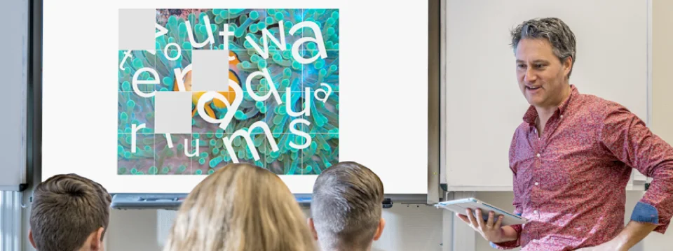 Een leraar staat met een tablet voor de klas, naast een digibord.